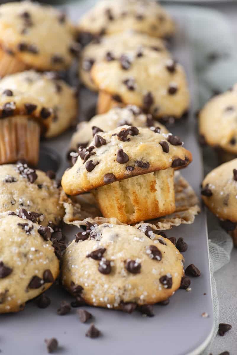 A close-up of freshly baked chocolate chip muffins in a muffin tin. One muffin is slightly raised with its paper liner peeled back, showing a golden, fluffy texture and chocolate chips sprinkled on top.