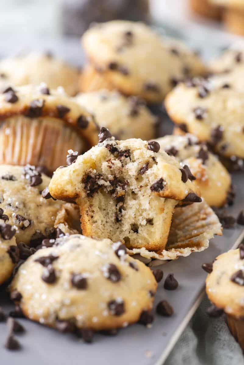 A close-up of several chocolate chip muffins, with one muffin broken in half to reveal its soft, moist interior. Chocolate chips are scattered on top of the muffins and on the surface around them.