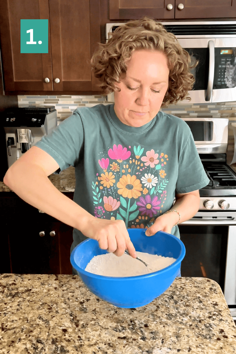 A woman with curly hair mixes flour in a blue bowl with a spoon in a kitchen. She wears a green t-shirt with colorful flowers, and there is a “1.” box in the upper left corner.