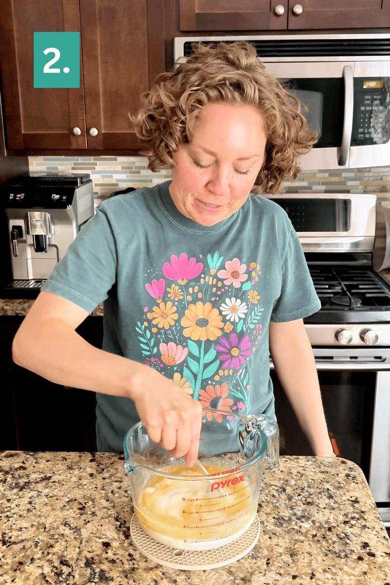 A person with curly hair wearing a teal shirt with colorful flowers whisks ingredients in a glass measuring cup on a kitchen counter. A green box with the number 2 is in the upper left corner.