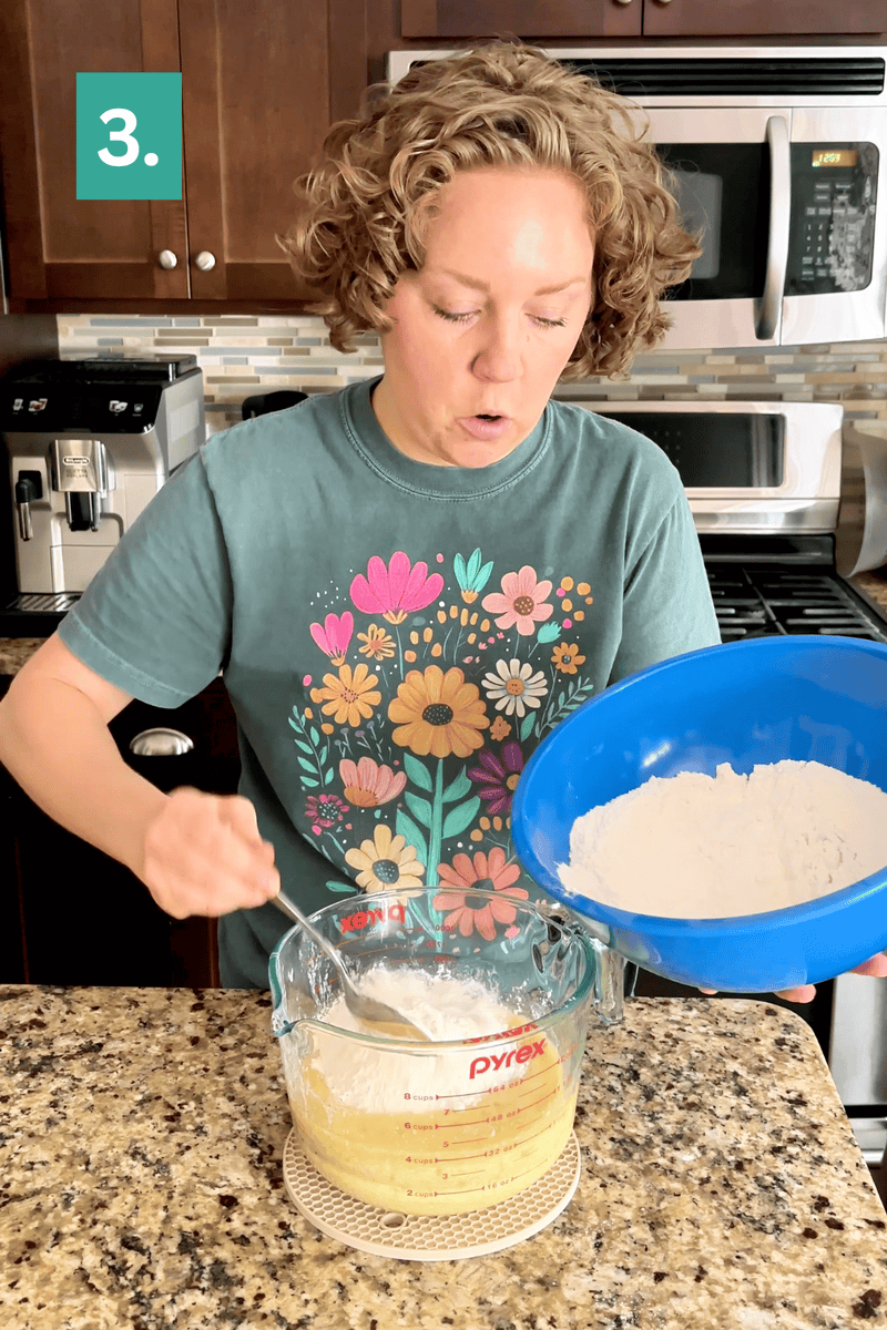 A person with curly hair adds flour from a blue bowl into a glass measuring cup on a kitchen counter. The person wears a green shirt with colorful flowers and stands in a modern kitchen. A green box labeled 3. is in the corner.