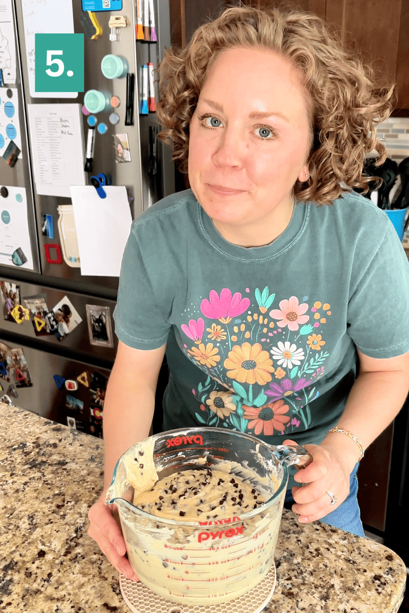 A woman with curly hair, wearing a green floral t-shirt, stands in a kitchen holding a Pyrex measuring cup filled with chocolate chip cookie dough. A 5. label appears in the top left corner.