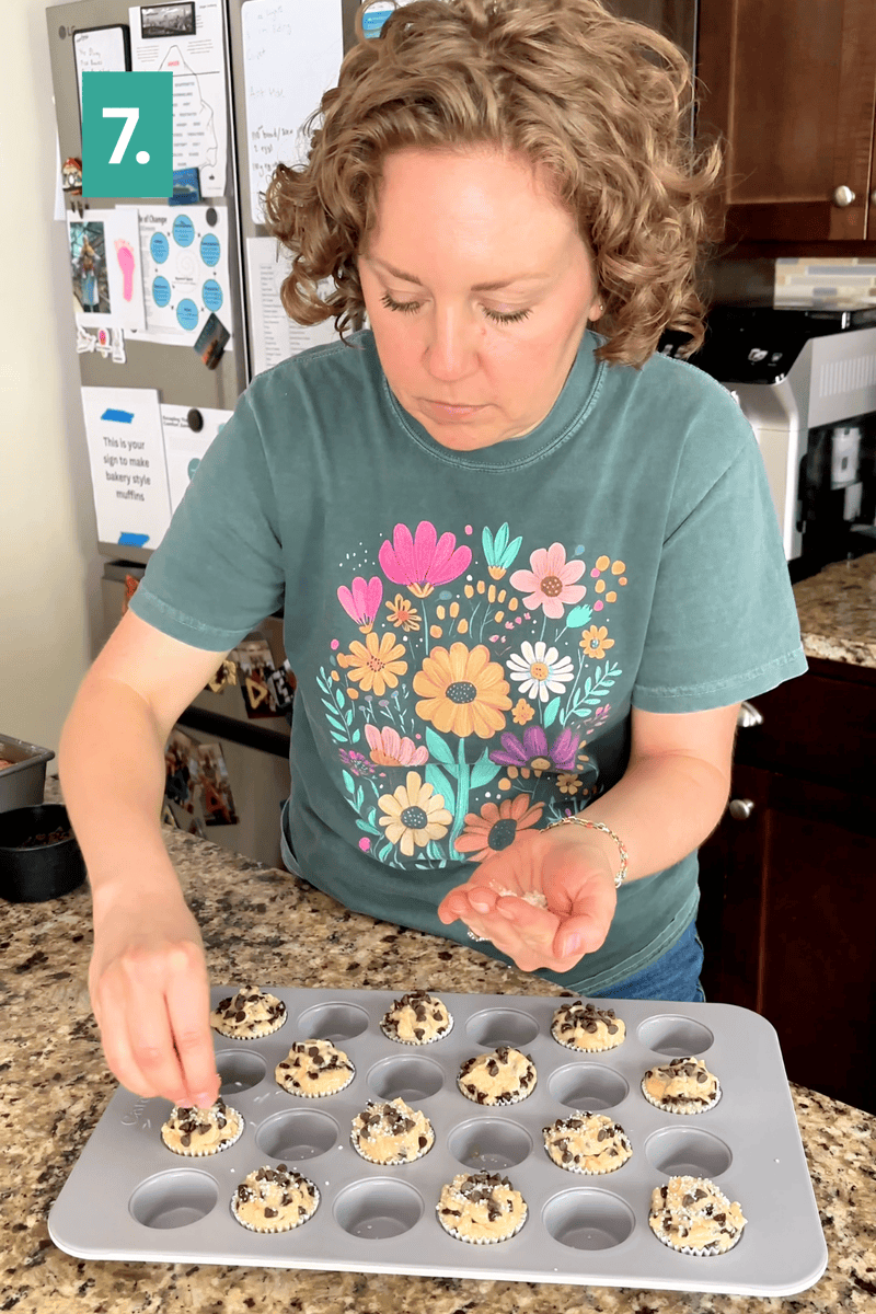 A person in a floral shirt adds toppings to muffin batter in a muffin tin on a granite kitchen counter. A 7. label is in the top left corner, and a refrigerator with notes is visible in the background.