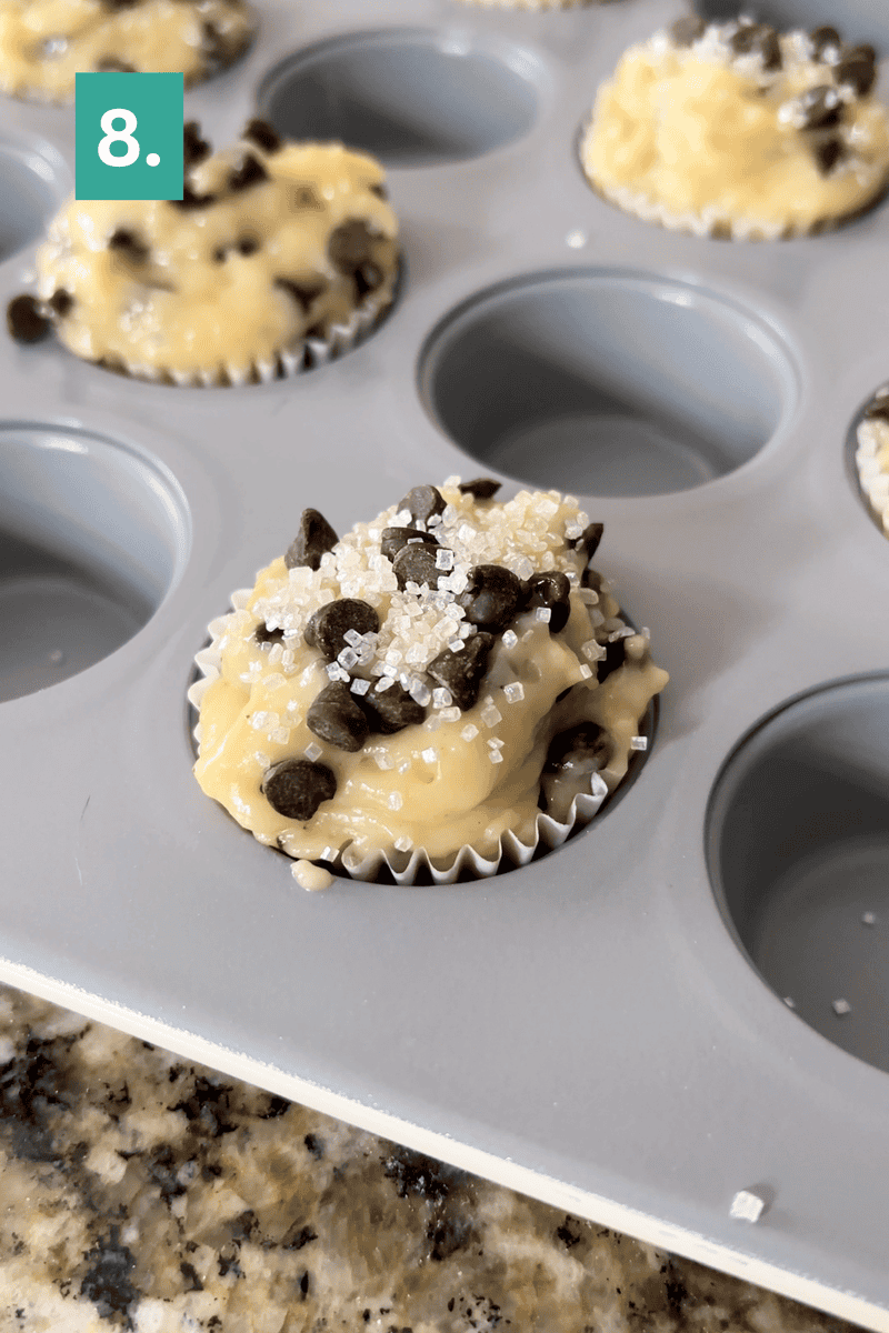 A muffin tray with muffin batter in paper liners, topped with chocolate chips and coarse sugar crystals. A green box with the number 8 is in the top left corner of the image.