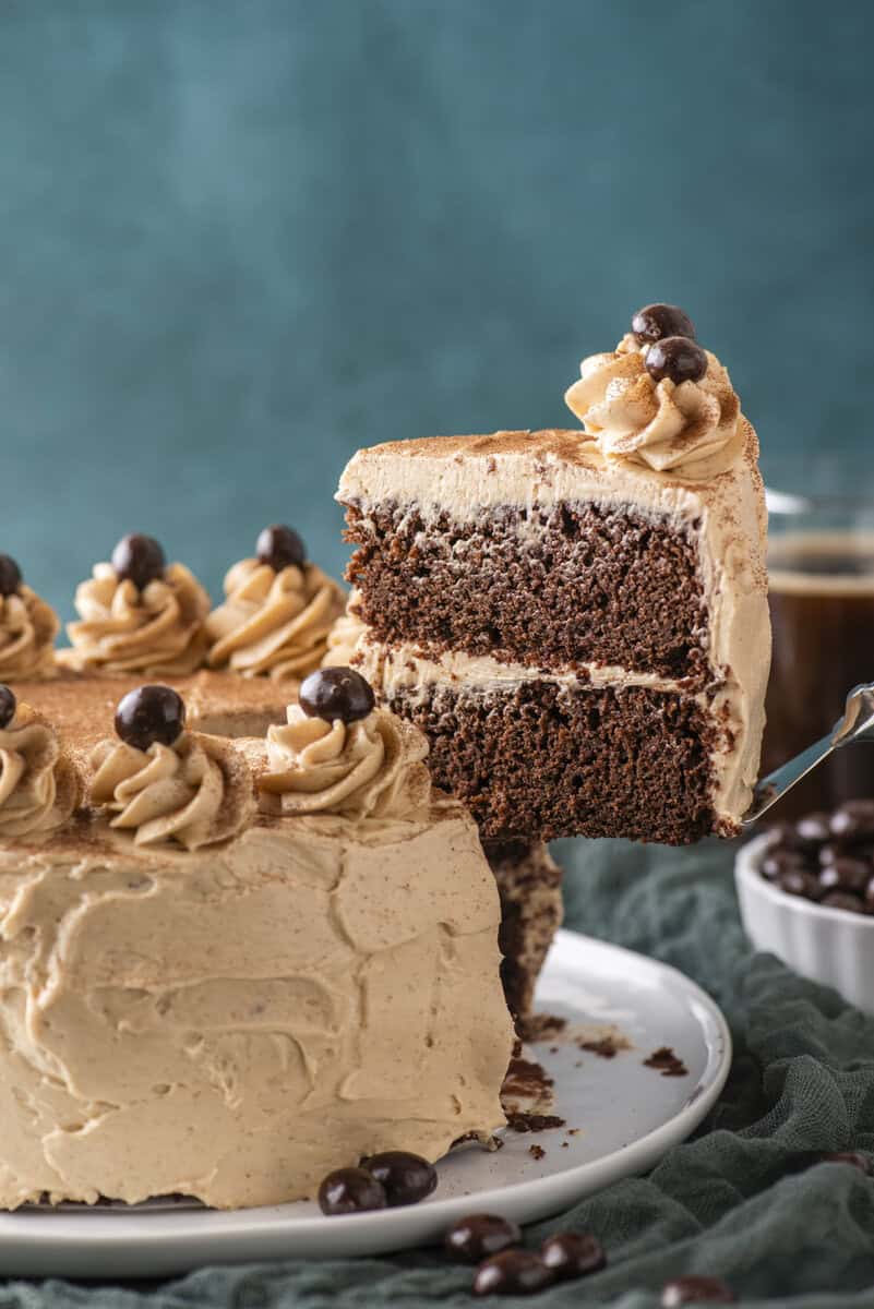 A slice of chocolate layer cake with light brown frosting is being lifted from a decorated cake, topped with swirls of frosting and dark round candies. A cup and a bowl of similar candies are in the background.