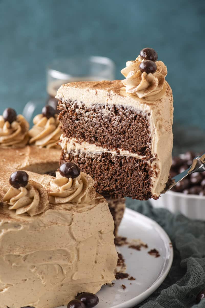 A slice of two-layer chocolate cake with light brown frosting and chocolate decorations is being lifted from a frosted cake on a white plate.