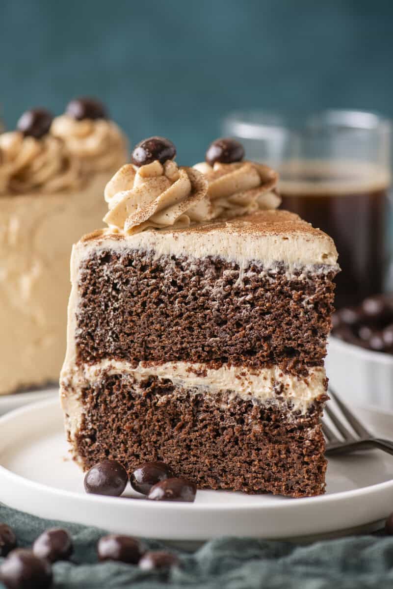 A slice of chocolate layer cake with light brown frosting and chocolate-covered coffee beans on top, served on a white plate with a fork. Two drinks and more cake are blurred in the background.