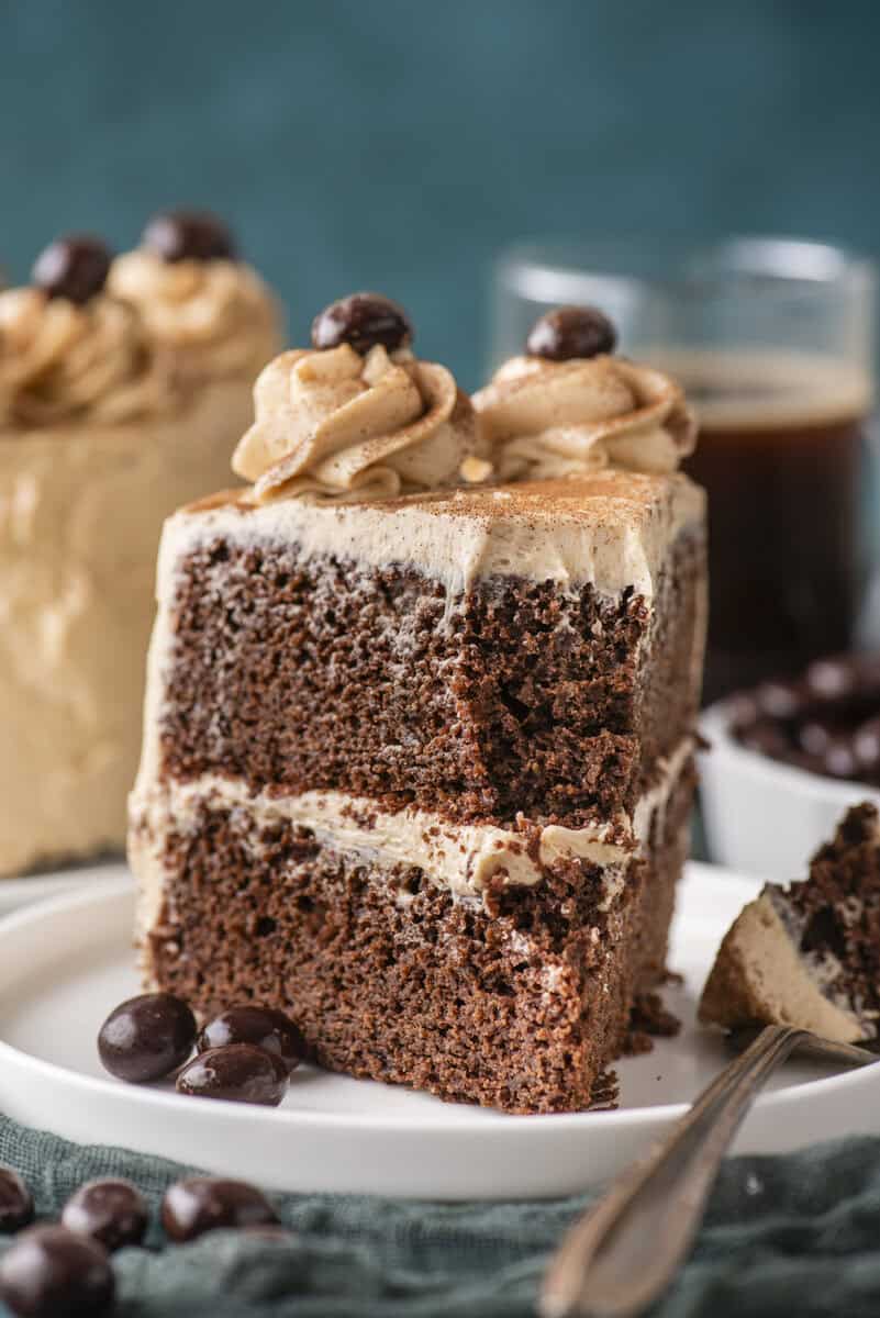 A slice of chocolate cake with creamy frosting, topped with swirls and chocolate-coated coffee beans, on a white plate with a fork. Blurred coffee and more cake are in the background.