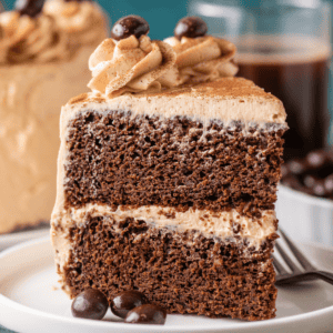 A slice of chocolate layer cake with light brown frosting, topped with piped swirls and chocolate-covered coffee beans, sits on a white plate with a fork, with more cake and coffee in the background.