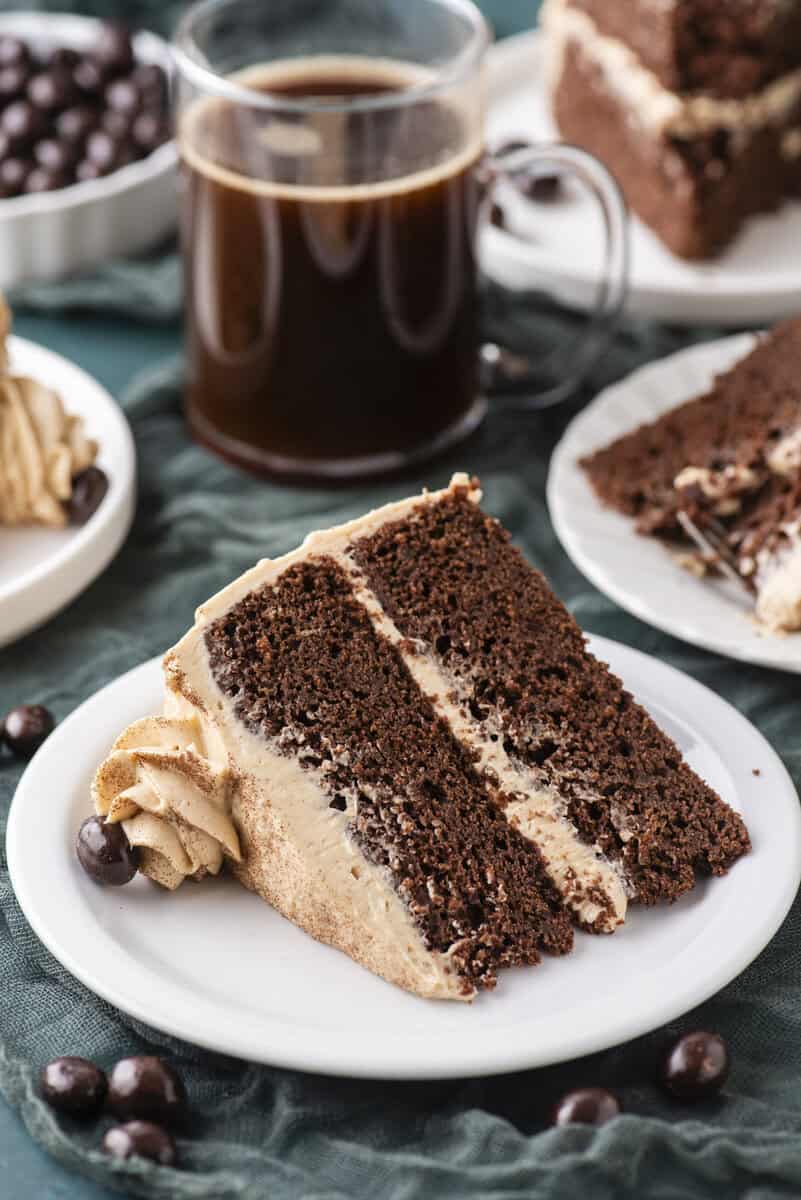 A slice of chocolate layer cake with light brown frosting on a white plate, next to a glass mug of black coffee and chocolate-covered coffee beans. More cake slices are visible in the background.