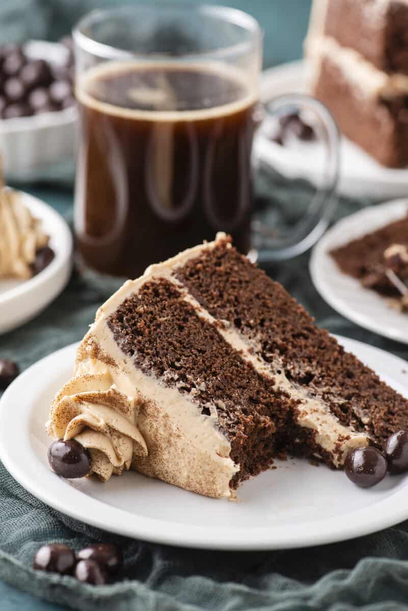 A slice of chocolate layer cake with mocha frosting sits on a white plate, with chocolate-covered coffee beans beside it. A clear mug of black coffee and more cake slices are in the background.