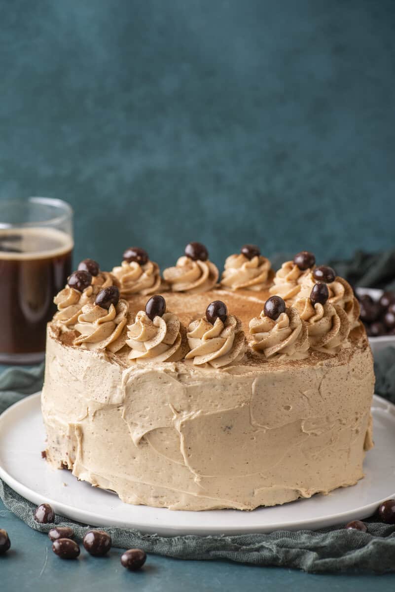Round cake with light brown frosting, decorated with swirls of frosting and chocolate-covered coffee beans on top, sits on a white plate. There is a glass of coffee and scattered beans nearby, set against a blue background.