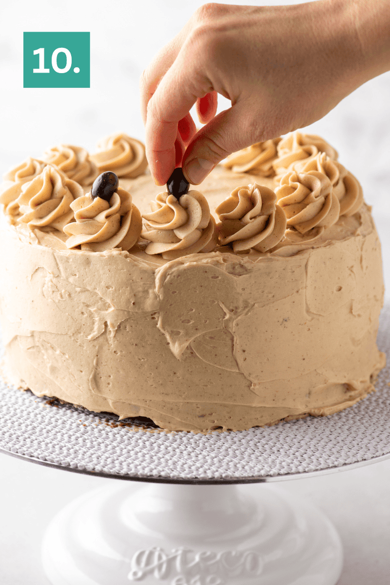 A hand places a coffee bean on top of a swirl of frosting on a round cake with light brown icing, displayed on a white cake stand. A green box with the number 10 appears in the upper left corner.