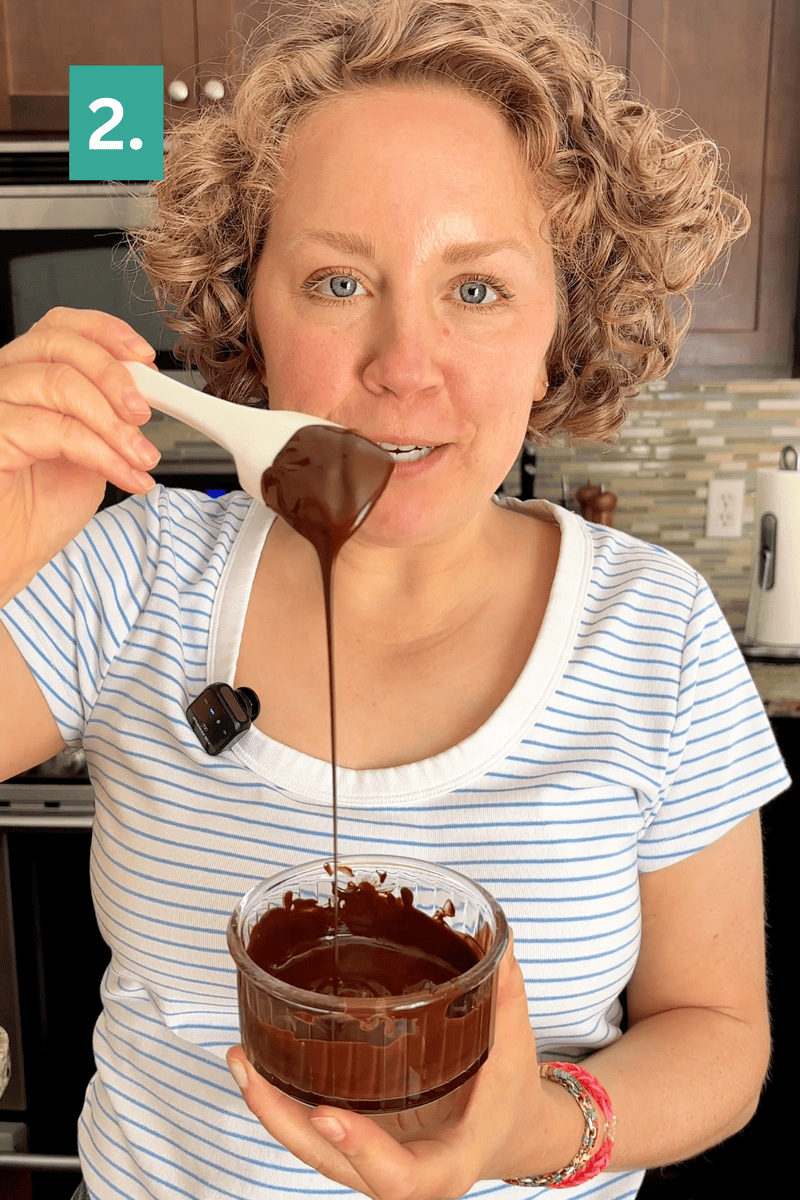 A woman in a striped shirt holds a glass bowl of melted chocolate and lifts some with a spoon. There is a 2. label in the top left corner. She stands in a kitchen with brown cabinets and a tiled backsplash.