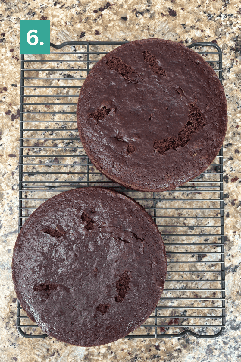 Two round chocolate cake layers cool on a wire rack on a granite countertop. The top layer has some cracks and holes. A green box with the number 6 is in the top left corner.