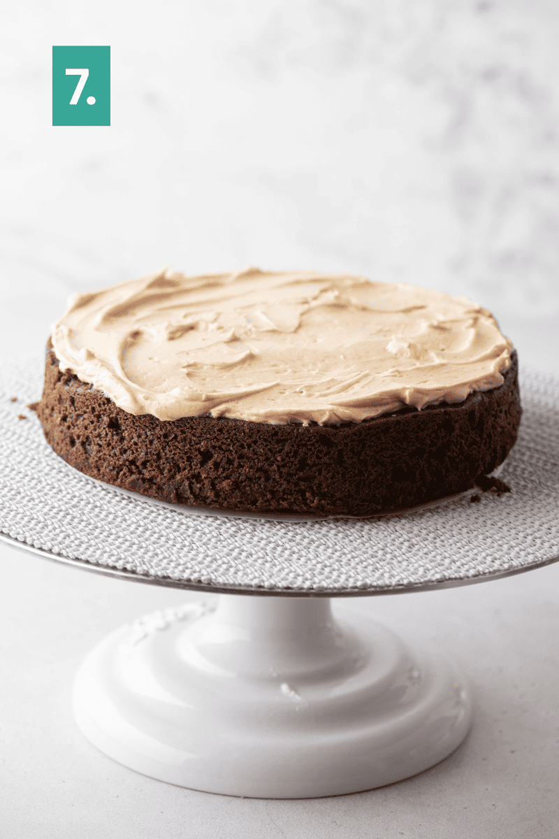 A round chocolate cake with light brown frosting sits on a white cake stand. There is a green square with the number 7 in the upper left corner.