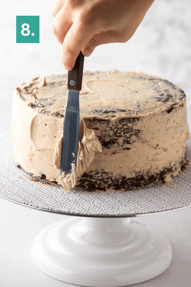 A hand uses a spatula to spread brown frosting on a round cake placed on a white cake stand. A green box with the number 8 appears in the top left corner.