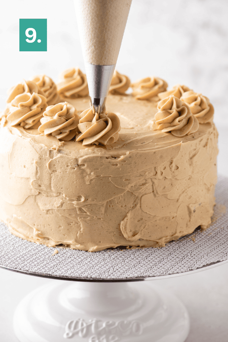 A piping bag adds swirls of light brown frosting to the top of a round cake on a white stand, with a green box showing the number 9 in the corner.