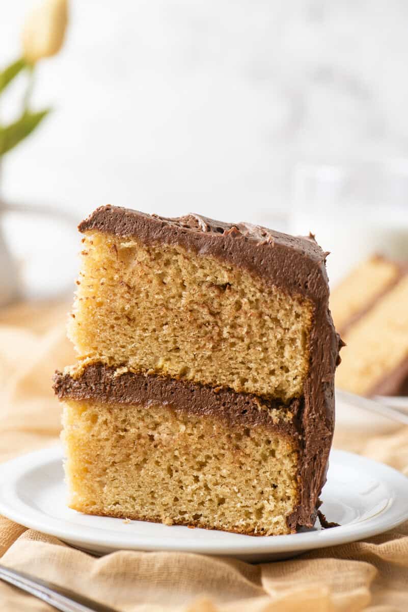 A close-up of a slice of yellow cake with chocolate frosting, stacked in two layers, served on a white plate with a blurred background.