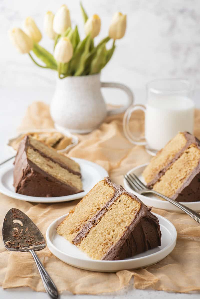 Three slices of yellow cake with chocolate frosting on white plates, forks beside them, with a white mug of milk and a vase of yellow tulips in the background on a peach-colored cloth.