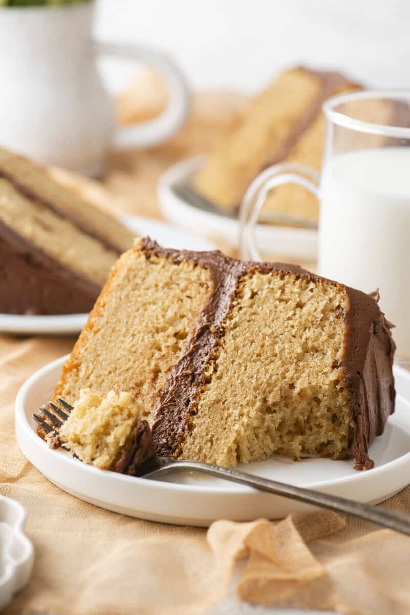 A slice of yellow cake with chocolate frosting sits on a white plate with a fork. A glass of milk and more cake slices are in the background.