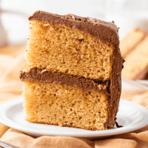 A slice of yellow cake with chocolate frosting between the layers and on top, served on a white plate with a blurred background.
