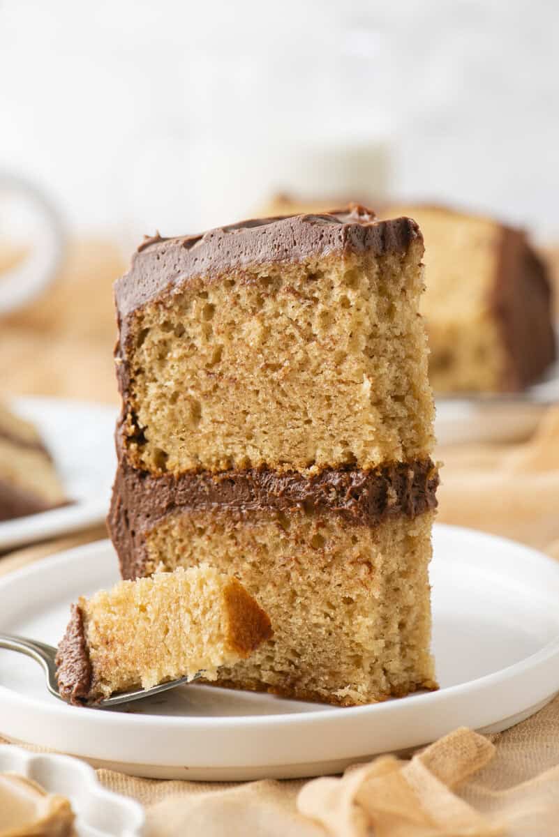 A slice of two-layer yellow cake with chocolate frosting sits on a white plate. A fork holds a bite-sized piece in front of the cake. The background is softly blurred.