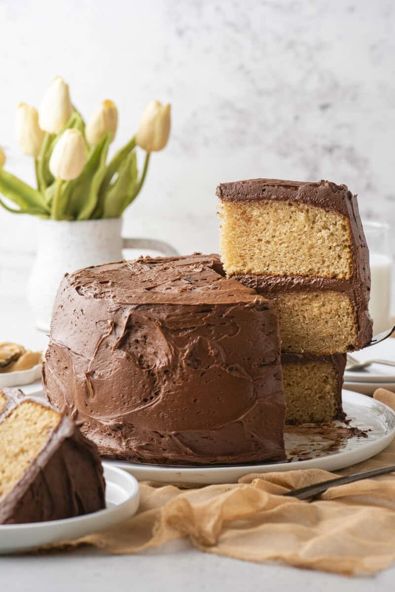 A two-layer yellow cake with chocolate frosting sits on a plate, with one slice lifted up. A vase of yellow tulips and a glass of milk are in the background.