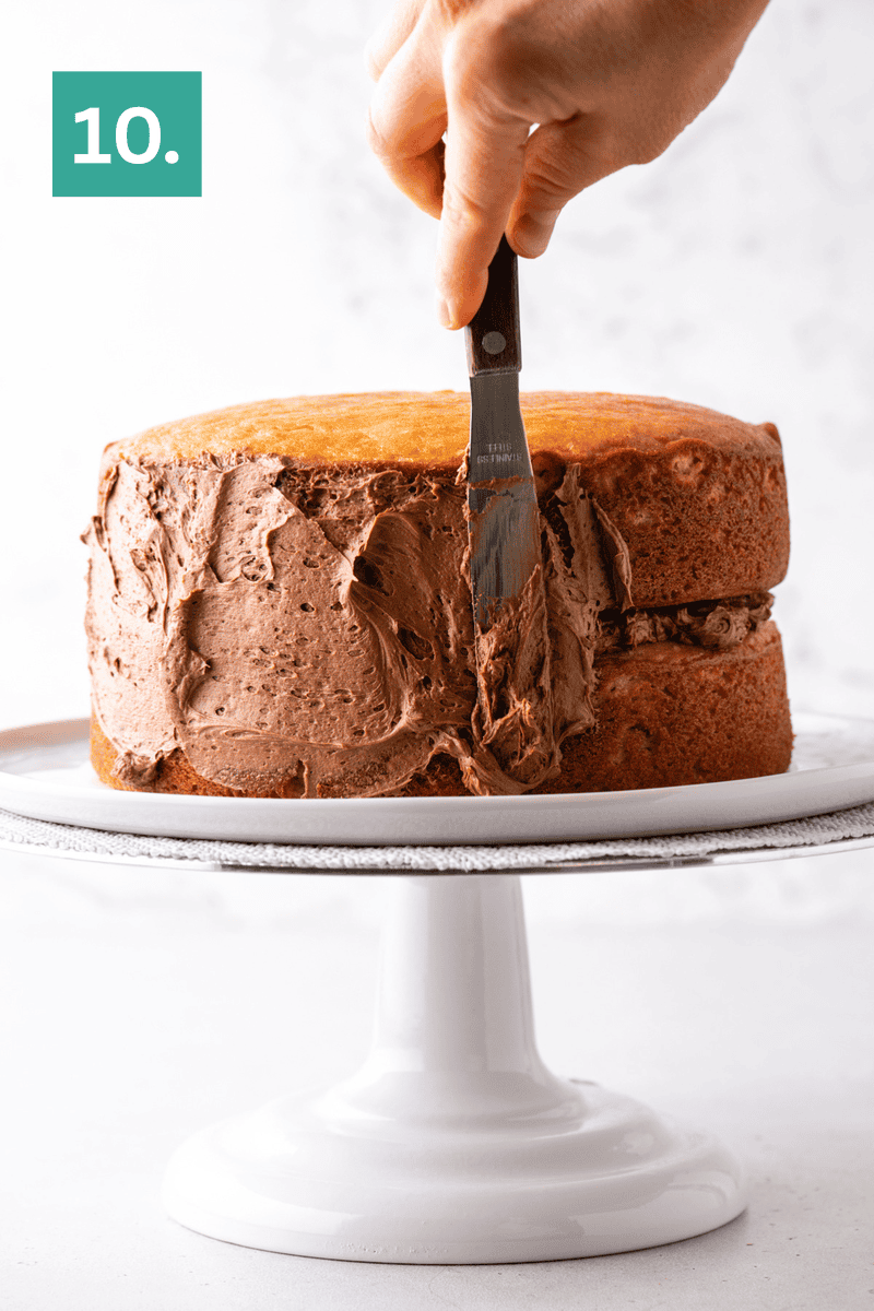 A hand uses a spatula to spread chocolate frosting on the side of a two-layer yellow cake, placed on a white cake stand. A green box with the number 10 is in the upper left corner.