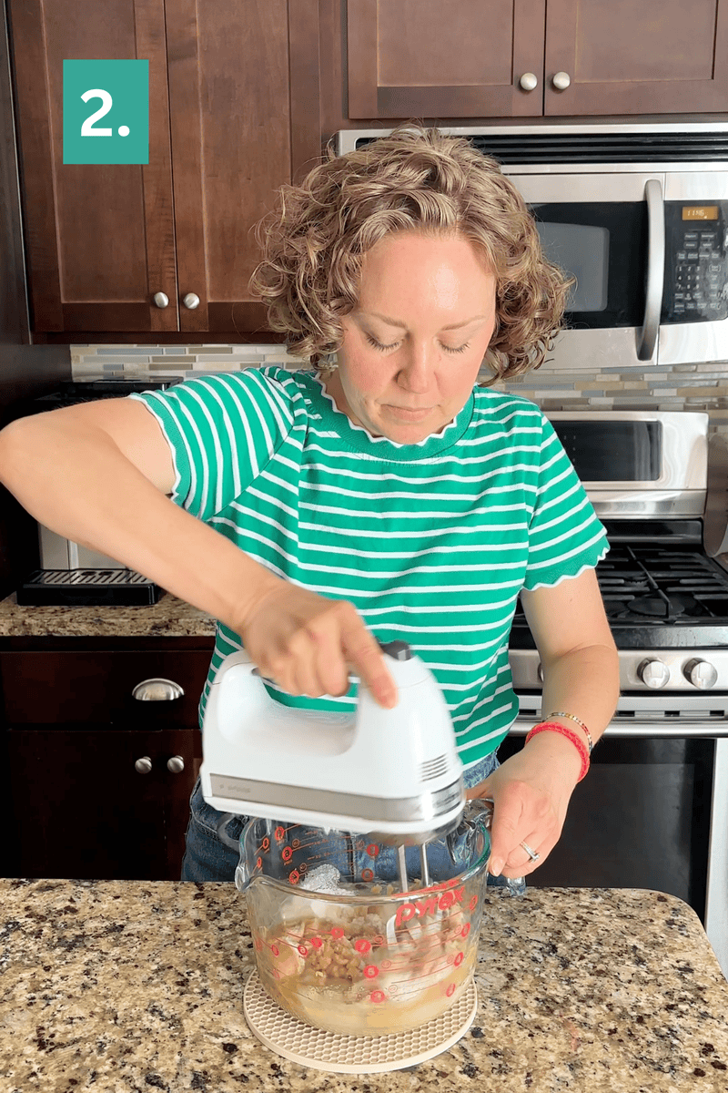 A person in a green striped shirt uses a hand mixer in a glass bowl in a kitchen. A stove, microwave, and wooden cabinets are visible in the background. A green box with the number 2 is in the top left corner.