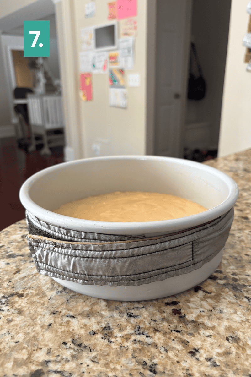 A bowl with batter sits on a granite counter, wrapped in a silver cloth strip. Colorful papers are taped to a wall in the background. A teal box with the number 7 is in the top left corner.