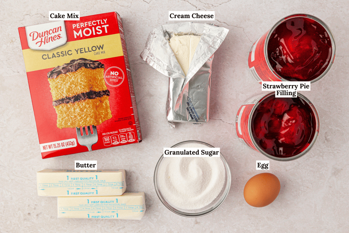 Labeled ingredients for a dessert: yellow cake mix, cream cheese, strawberry pie filling, two butter sticks, granulated sugar in a bowl, and an egg arranged on a light surface.