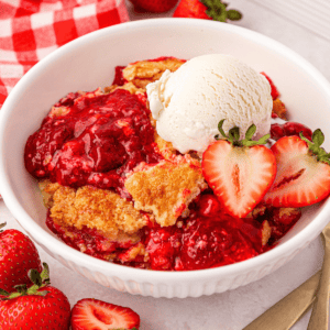 A white bowl filled with strawberry cobbler topped with a scoop of vanilla ice cream and fresh strawberry halves, surrounded by whole and sliced strawberries.