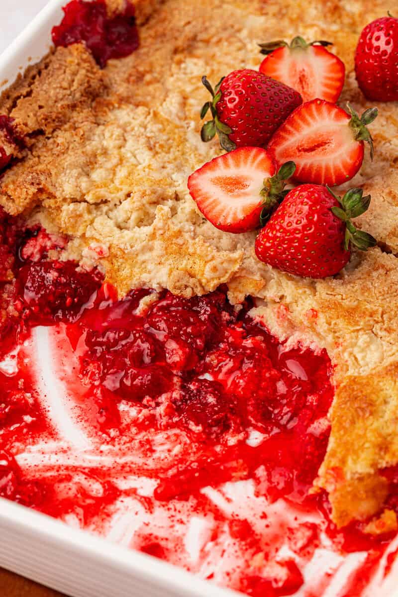A partially eaten berry cobbler in a white dish, topped with fresh strawberries and golden, crumbly crust. Bright red berry filling is visible where pieces have been served.