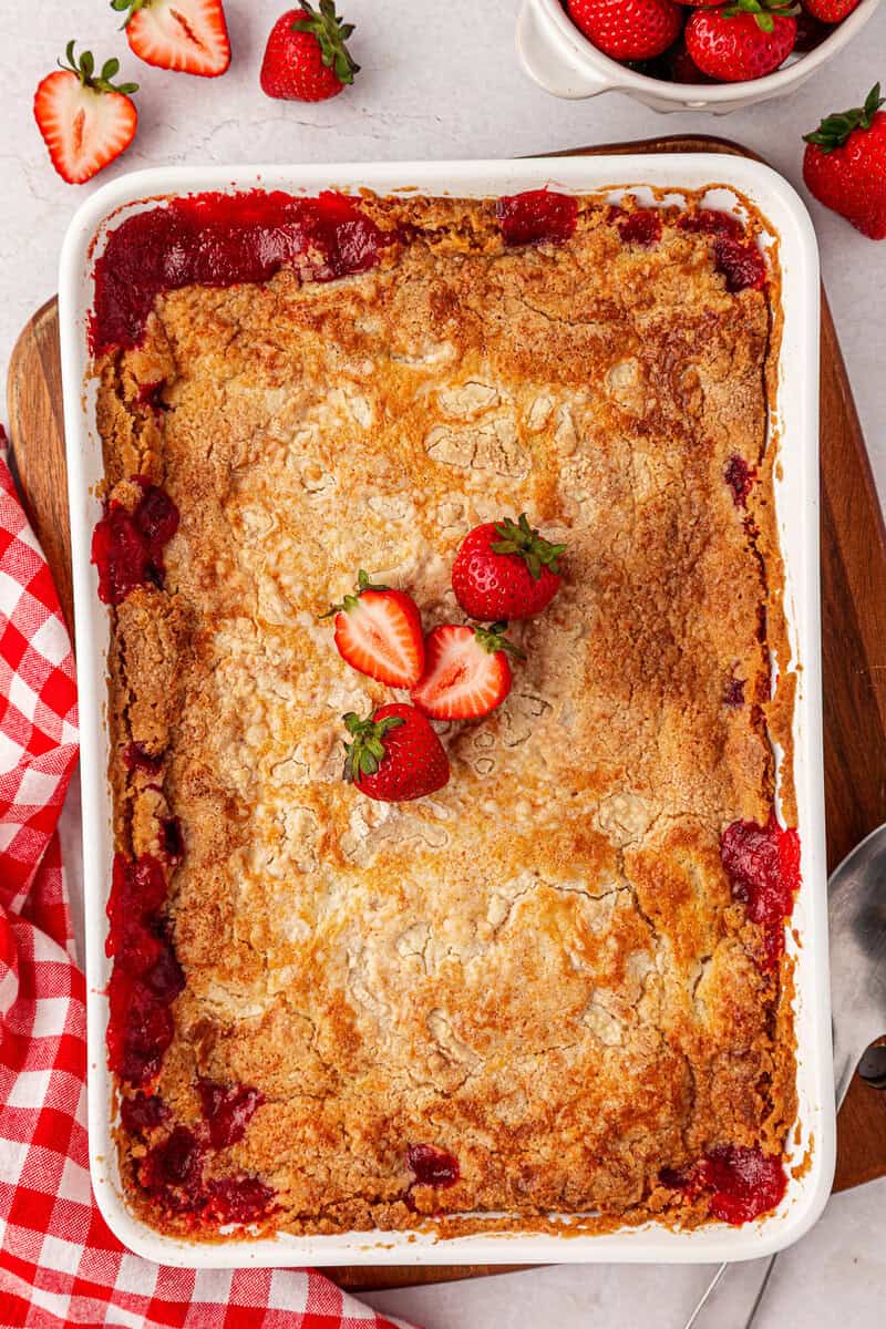A baked strawberry cobbler in a white dish, topped with three fresh strawberries, sits next to a red checkered cloth and strawberries on a white surface.
