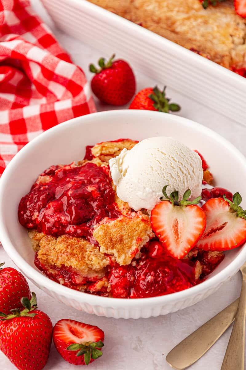 A bowl of strawberry cobbler topped with a scoop of vanilla ice cream and sliced strawberries, with fresh strawberries and a red checkered napkin nearby.