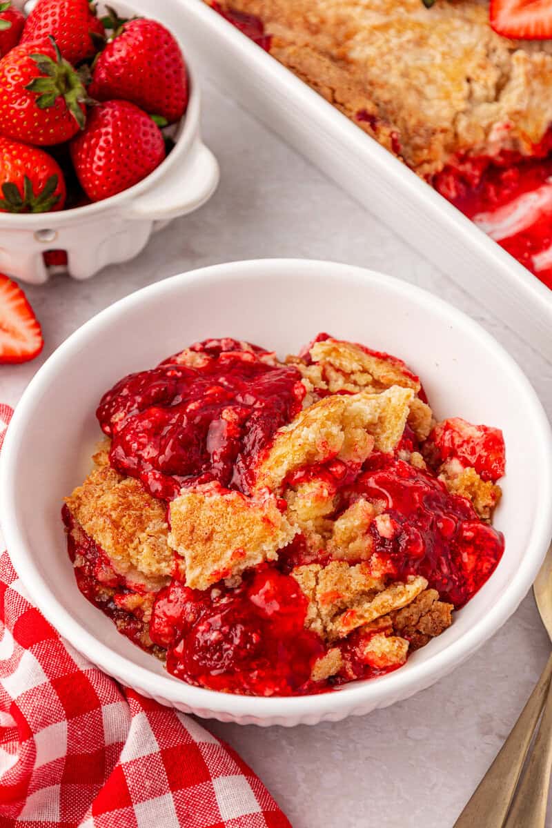 A white bowl filled with strawberry cobbler sits next to a dish of fresh strawberries and a red checkered napkin on a white surface.