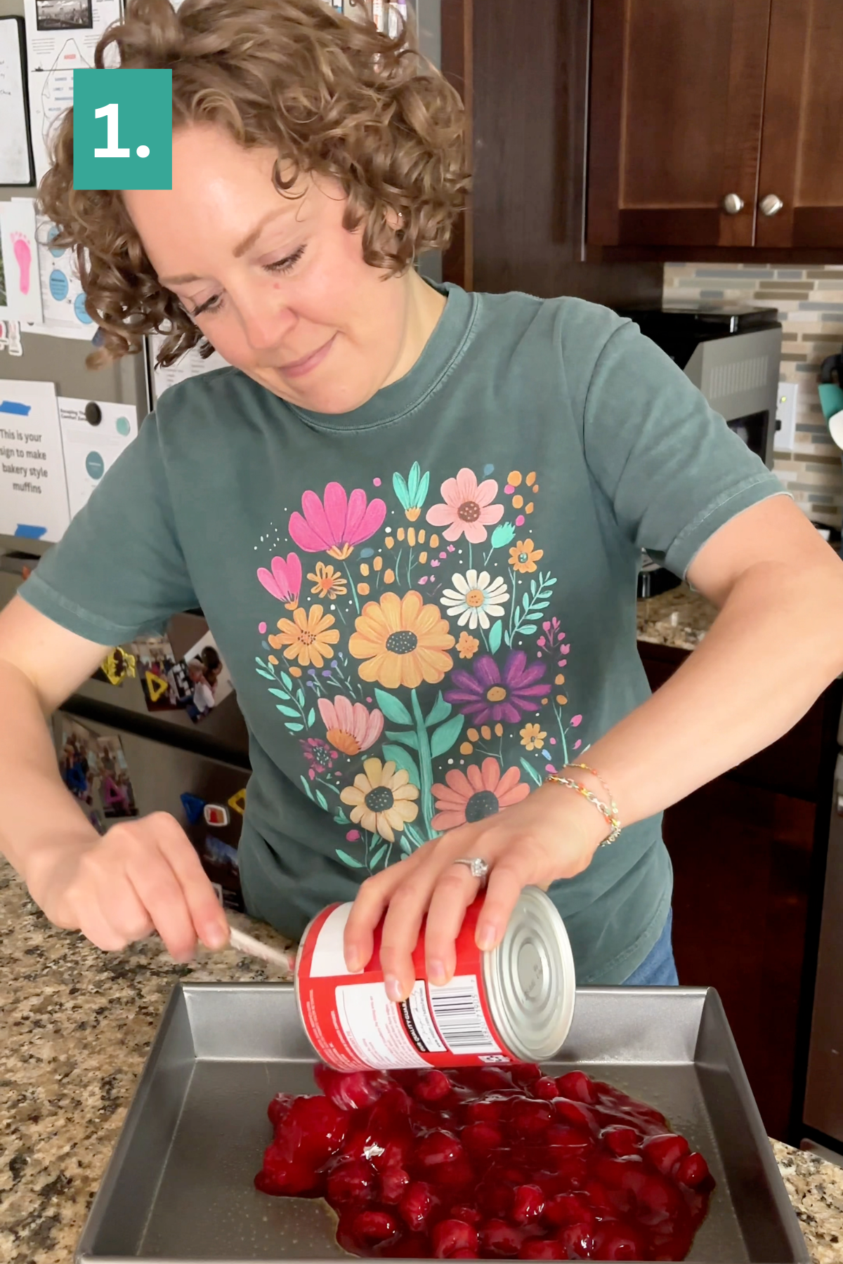 A woman with curly hair pours canned cherry filling into a baking pan on a kitchen counter, starting the first step of a Strawberry Cheesecake Dump Cake. She wears a green T-shirt with colorful flowers. A 1. appears in the corner, indicating the initial step of this delicious recipe.