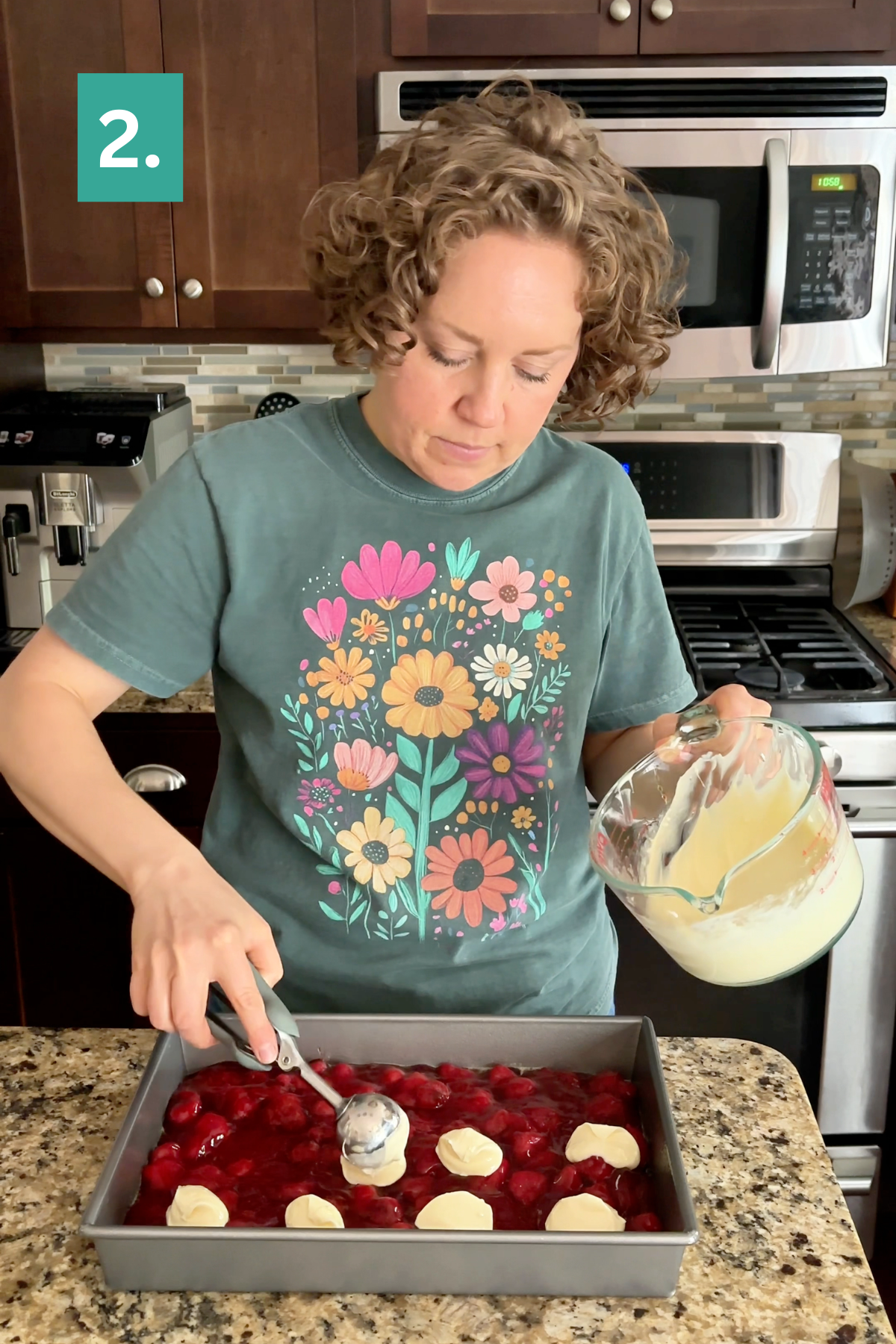 A person in a flower-print shirt scoops batter onto a Strawberry Cheesecake Dump Cake in a pan, standing in a kitchen with dark cabinets, a microwave, and a stove.
