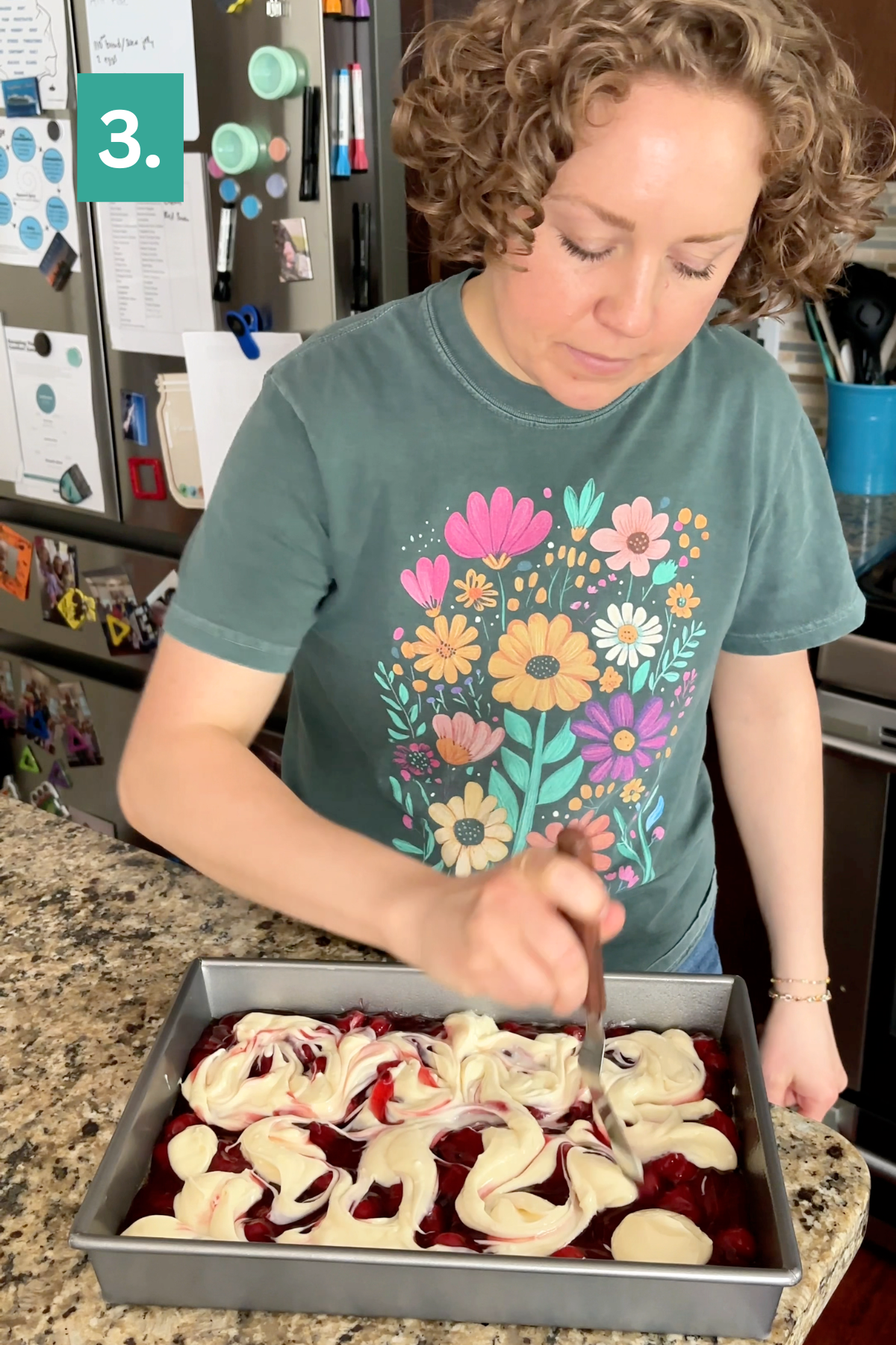 A person with curly hair swirls cream cheese into a vibrant red batter for a Strawberry Cheesecake Dump Cake in a cake pan on the kitchen counter. They wear a green floral shirt, and the kitchen background features magnets, notes, and utensils. A green 3. is visible in the top left corner.