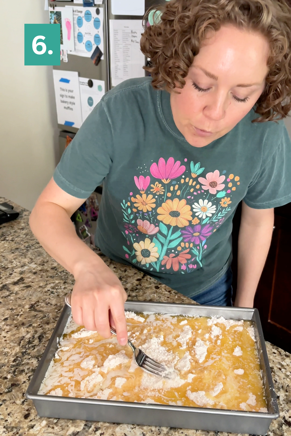 A woman with curly hair uses a fork to mix powdered ingredients into a yellow mixture in a baking pan, preparing what looks like a Strawberry Cheesecake Dump Cake, while standing at a granite countertop. A green box with the number 6 is in the upper left corner of the image.