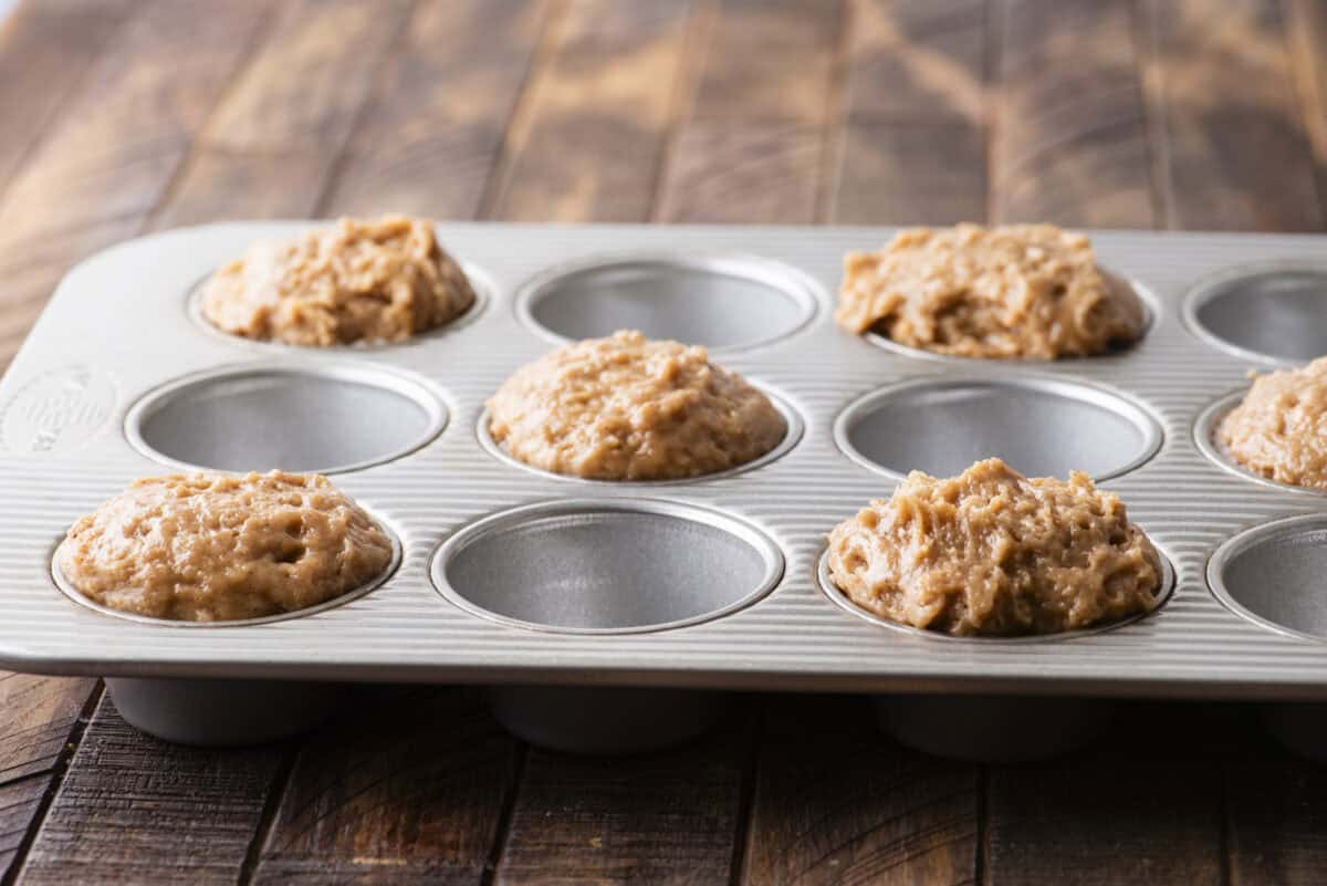 A muffin tin filled with several portions of raw muffin batter, ready to be baked, sits on a wooden surface.