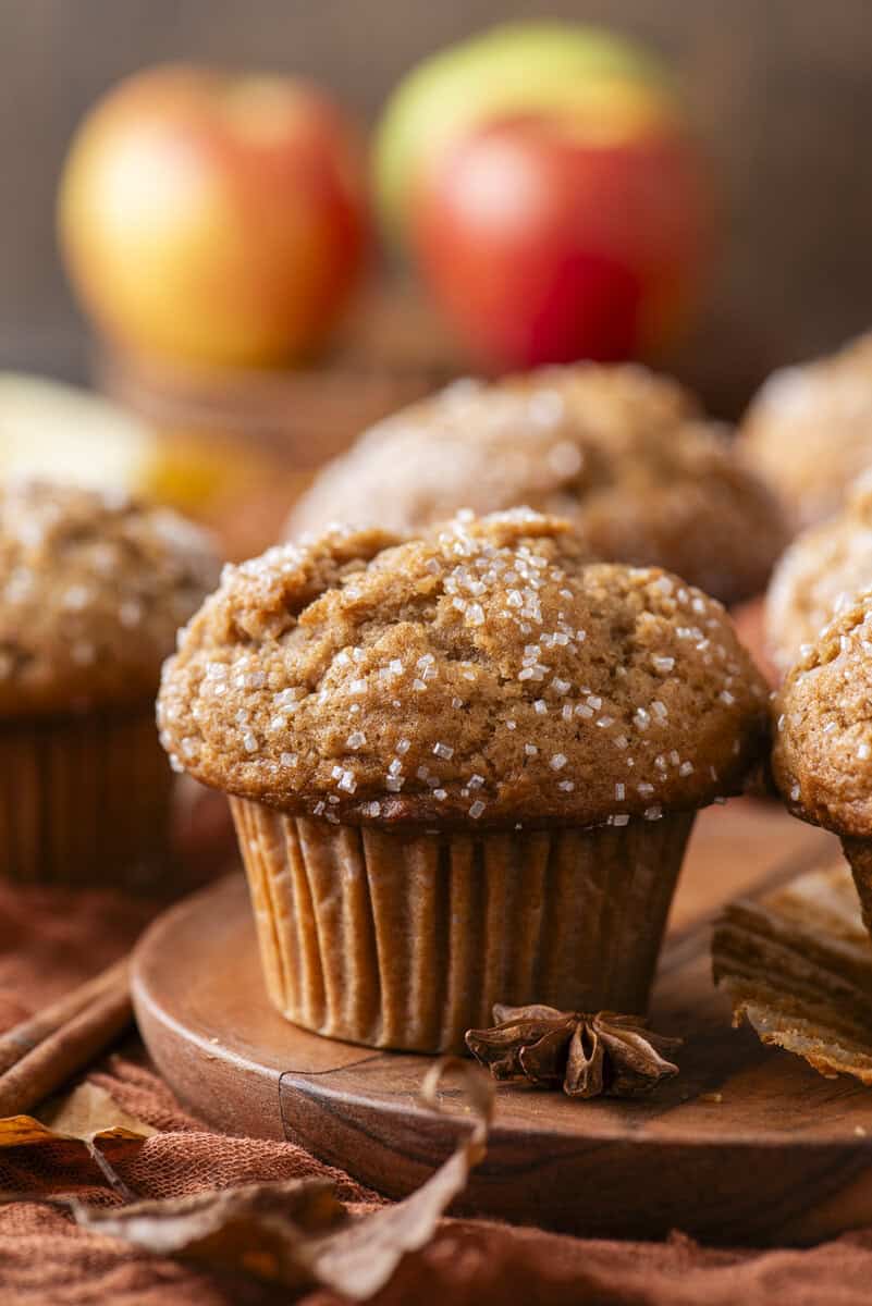 A close-up of a spiced muffin topped with coarse sugar sits on a wooden plate, with more muffins and blurred apples in the background. The scene includes warm autumn colors and whole star anise.