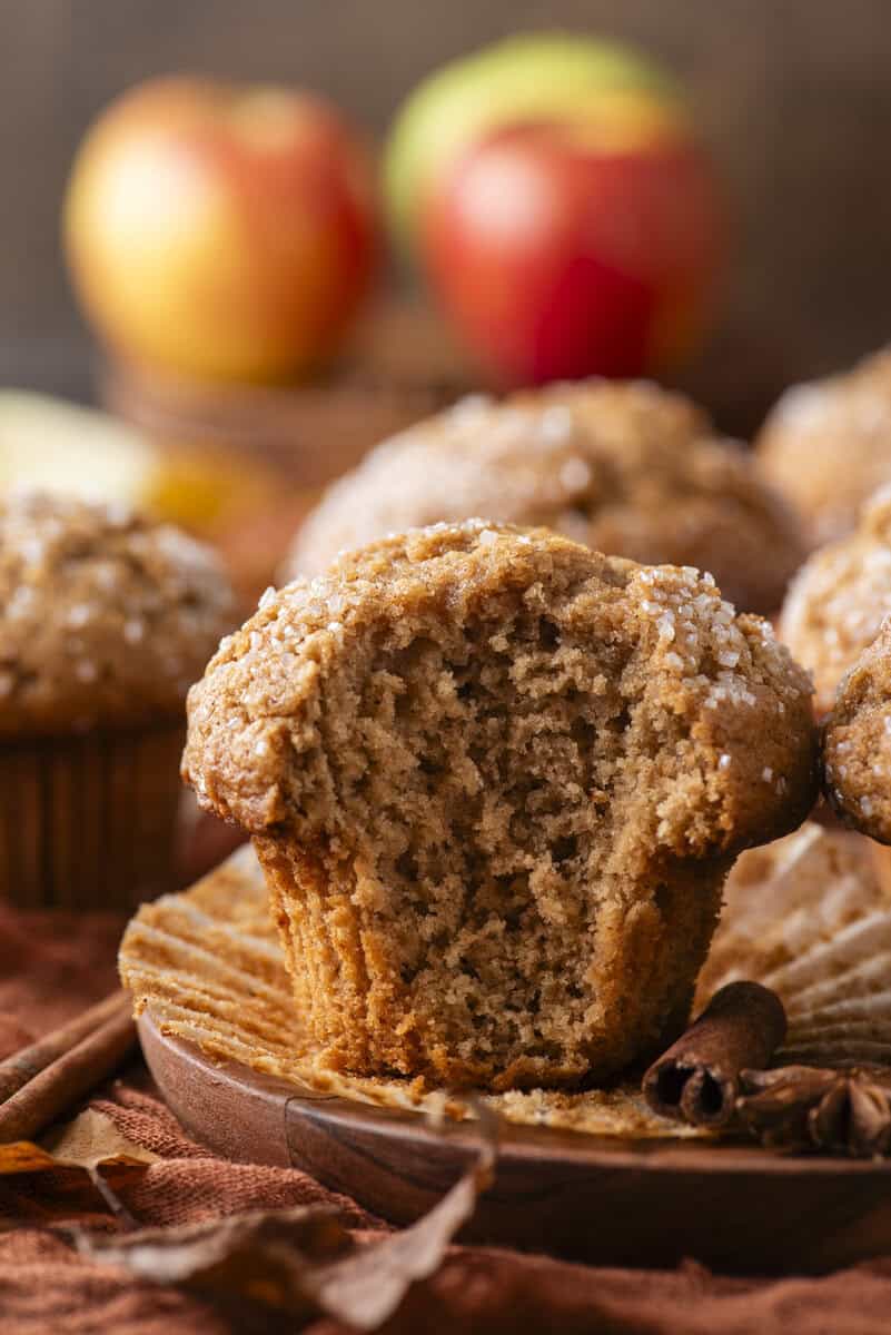 A cinnamon muffin with a bite taken out sits on a wooden plate, surrounded by more muffins, apples, and cinnamon sticks in the background.
