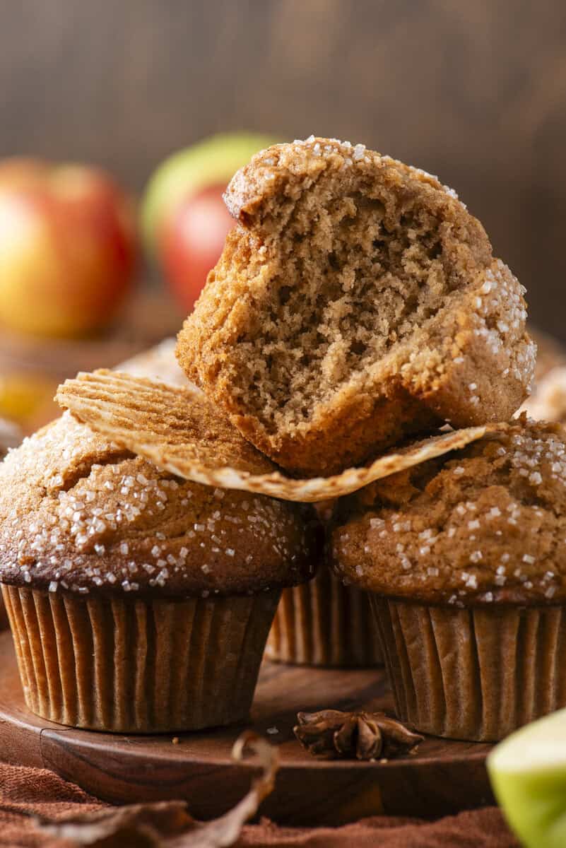 Three brown muffins sprinkled with coarse sugar sit on a wooden surface. One muffin, with a bite taken out, is resting on top of the others. Apples are blurred in the background.