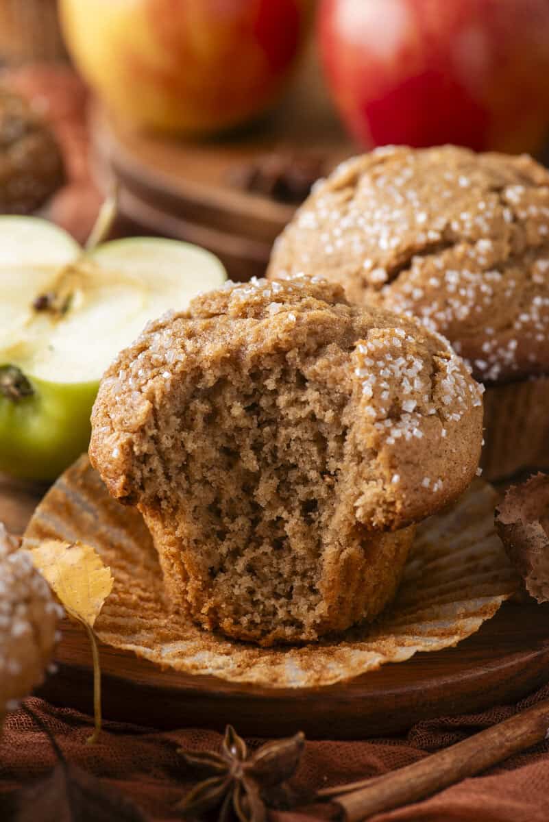 A spiced muffin with a bite taken out sits in a paper liner. It is topped with coarse sugar. Another muffin, apples, and autumn decorations are in the blurred background.