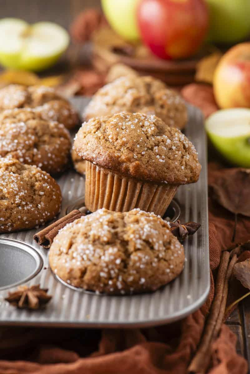 A muffin tin holds several apple muffins topped with coarse sugar, with apples and spices like cinnamon and star anise in the background, on a rustic surface.