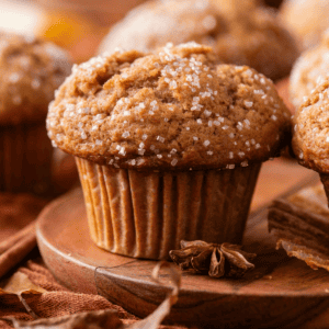 A close-up of a spiced muffin topped with coarse sugar crystals, sitting on a wooden plate with cinnamon sticks and star anise nearby. Other muffins are blurred in the background.
