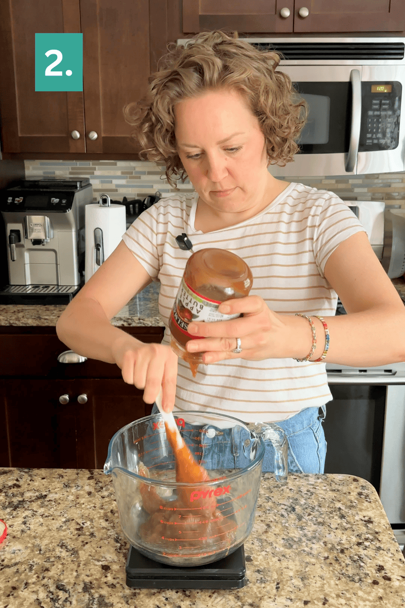 A woman in a striped shirt pours tomato sauce from a jar into a glass measuring cup, using a spatula, in a modern kitchen. A green box labeled 2. appears in the top left corner.