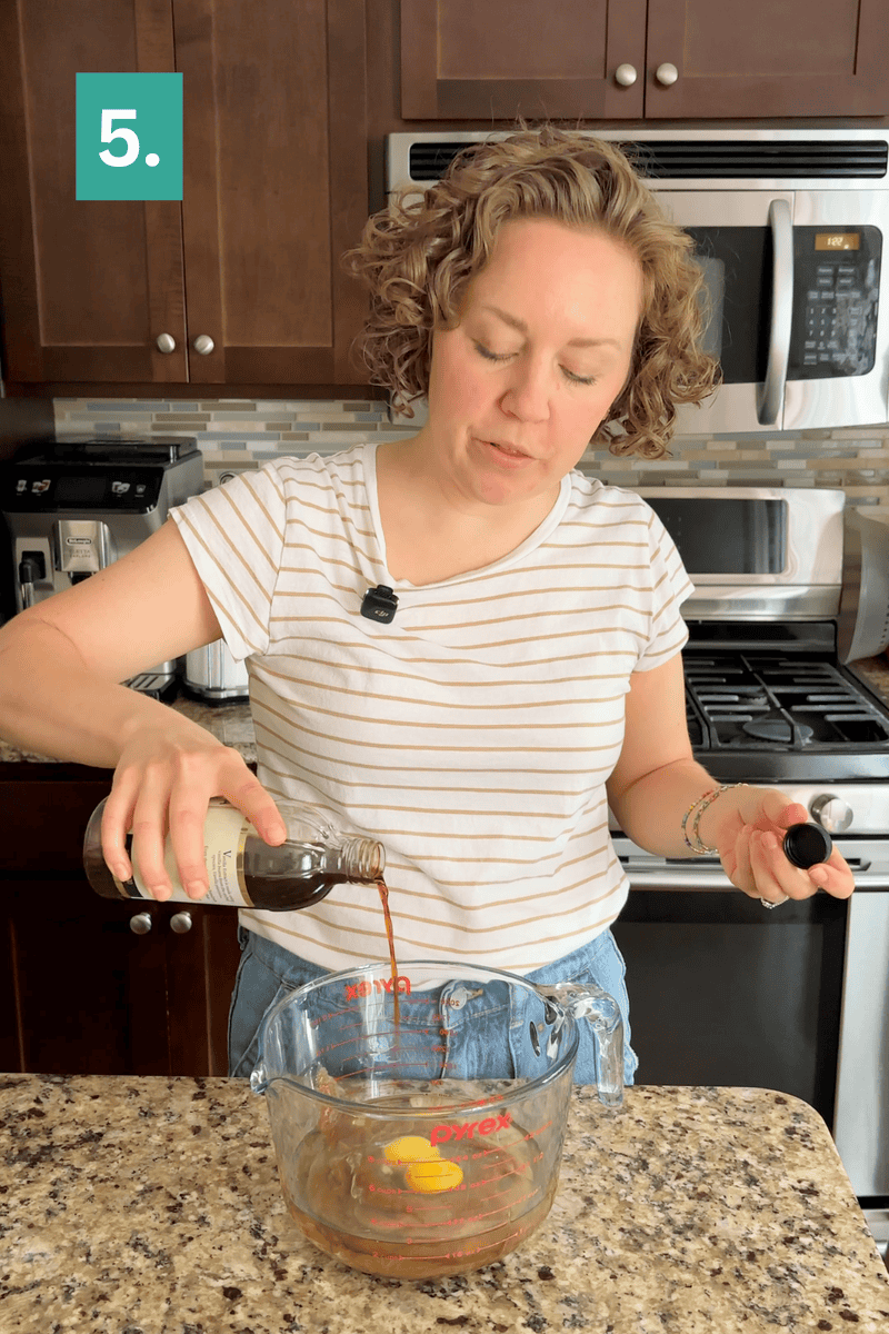 A woman pours liquid from a bottle into a measuring cup with eggs on a kitchen counter. A green square with 5. appears in the top left corner.